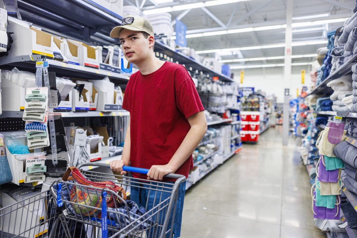 Young man with autism pushes shopping cart through store aisle. He wears red t-shirt and cap, focusing intently on surrounding shelves. Scene captures autistic individual managing sensory-rich retail environment, highlighting everyday challenges and independence of neurodivergent shoppers.
